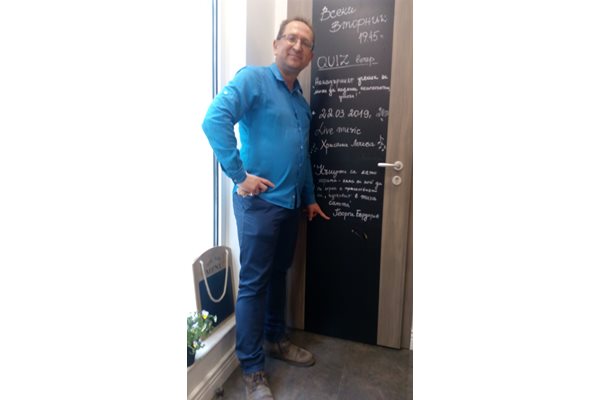 Георги Бърдаров пред вратата към тоалетната в бар "Каре арте" в София. На нея собствениците на заведението са изписали цитат от разказа "За петата ракия или колко е хубав животът". Писателят гордо сочи името си.