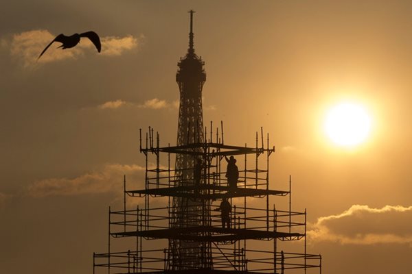 Силуети на работници на скеле пред Айфеловата кула, докато слънцето залязва над Париж.