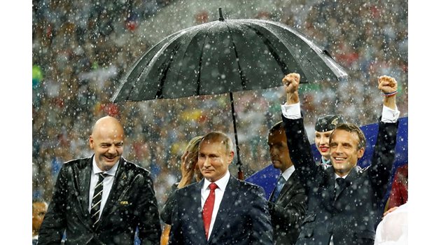 Служител държи чадър над главата на Владимир Путин, вдясно е Еманюел Макрон, вляво - Джани Инфантино.   СНИМКА: РОЙТЕРС