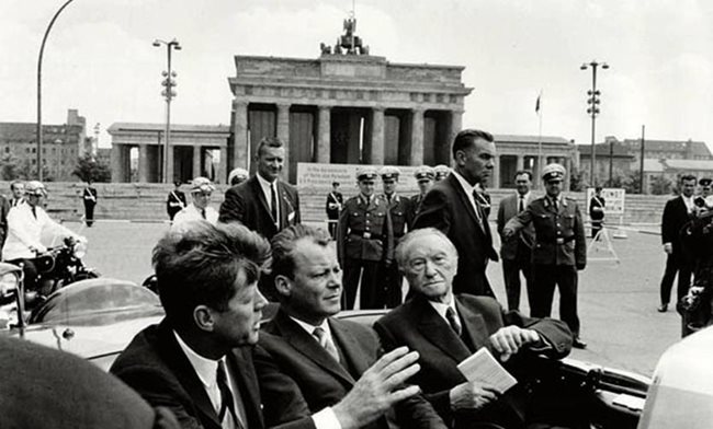 Президентът Джон Кенеди, кметът на Западен Берлин Вили Брант и канцлерът на Германия Конрад Аденауер в кола пред Бранденбургската врата през 1963 г. В Западен Берлин Кенеди изрича историческата фраза “Аз съм берлинчанин” на немски, без да знае езика.